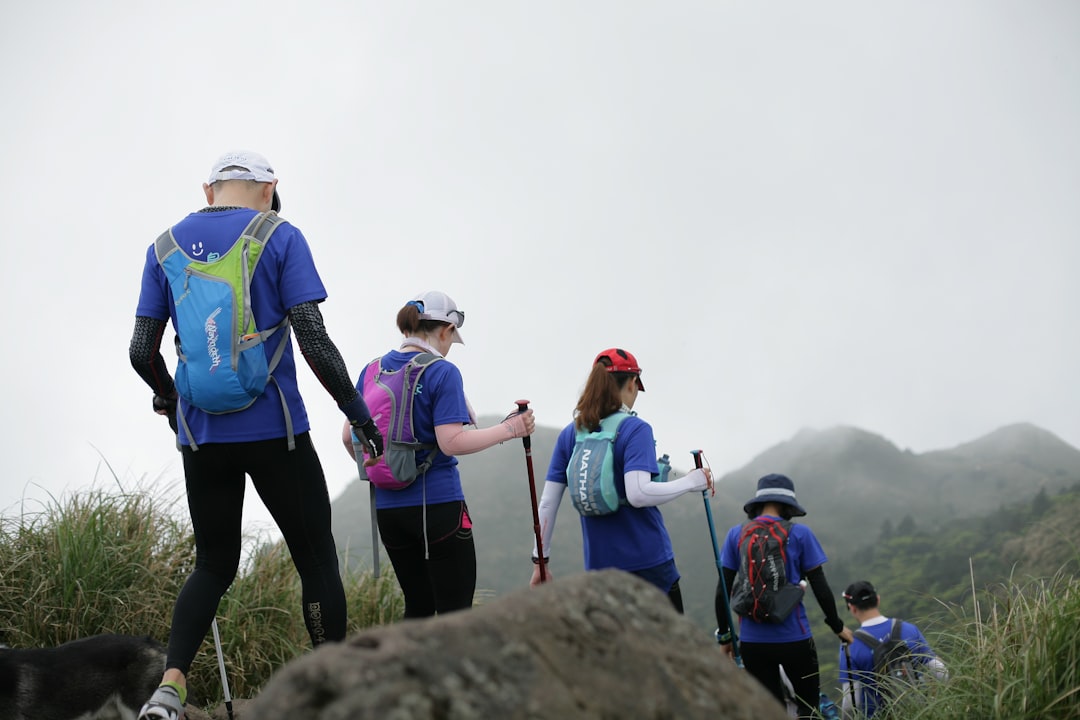 Das Bild zeigt eine Gruppe von Menschen, die bei nebligem Wetter in den Bergen wandern. Alle tragen blaue T-Shirts, Rucksäcke und Wanderstöcke. Die Gruppe bewegt sich einen Pfad entlang, der von Gras und Felsen gesäumt ist, und im Hintergrund sind bergige Landschaften zu sehen. Dieses Bild passt perfekt zum Abschnitt über Outdoor-Abenteuer im Blog-Beitrag und illustriert die Teamaktivität des Wanderns. Es unterstreicht die Aspekte der körperlichen Gesundheit, des Teamzusammenhalts und des gemeinsamen Erlebens von Natur und Abenteuer. Die gemeinsame Bewältigung einer Wanderung fördert die Zusammenarbeit und stärkt das Vertrauen unter den Teammitgliedern.